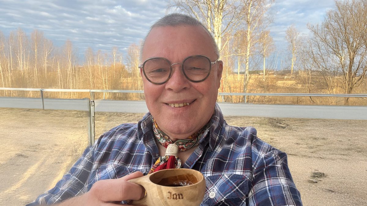 Person i rutig skjorta och scarf håller en träkopp med dryck utomhus på en plats med bar mark och träd i bakgrunden.