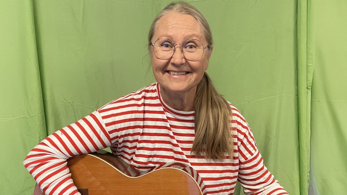 Kvinna med långt hår i hästsvans och randig tröja spelar akustisk gitarr framför en grön bakgrund.